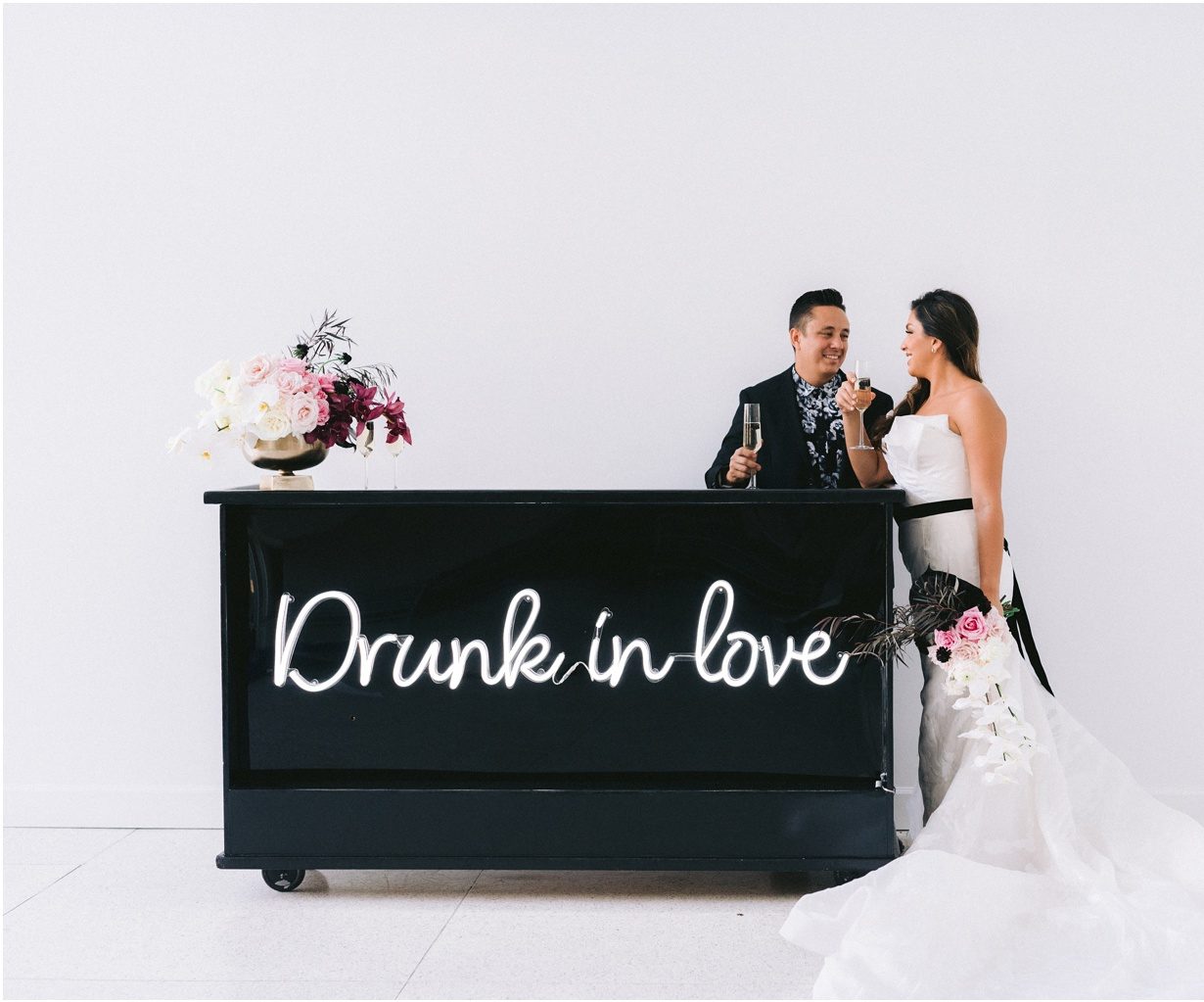 bride and groom standing in front of their wedding bar with a neon sign that says "drunk in love"